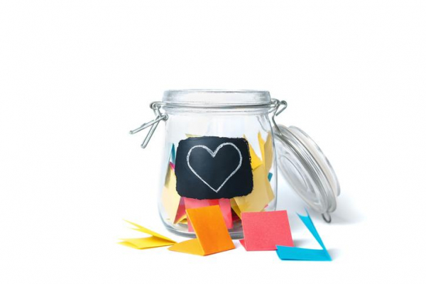 A clear jar with a heart label and colorful folded notes inside & scattered nearby against a white background 